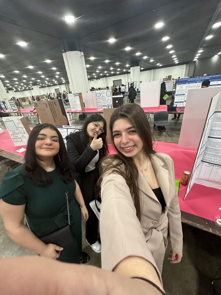 Three young ladies in a large hall full of science fair posters.