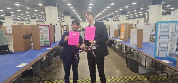 Two young men holding pink papers in a large hall full of science fair boards